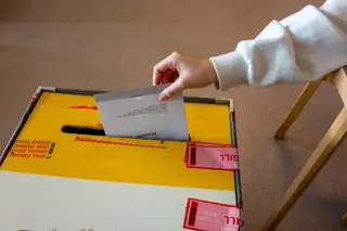 A hand dropping a sealed ballot envelope into a tamper-sealed ballot box at a polling station