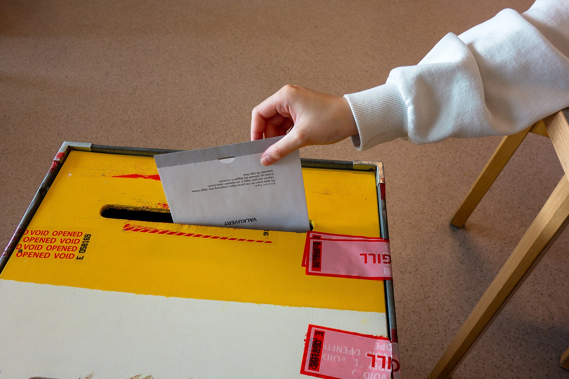 A hand dropping a sealed ballot envelope into a tamper-sealed ballot box at a polling station